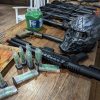 black airsoft guns lined up on a wooden counter with green pellets in a container and a black and grey protective mask