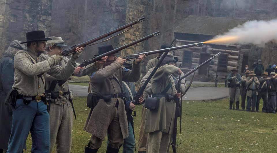 men in civil war uniforms shooting muskets in front of a wood cabin