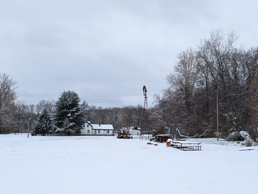 large snowy field with white farmhouse in background surrounded by barren trees